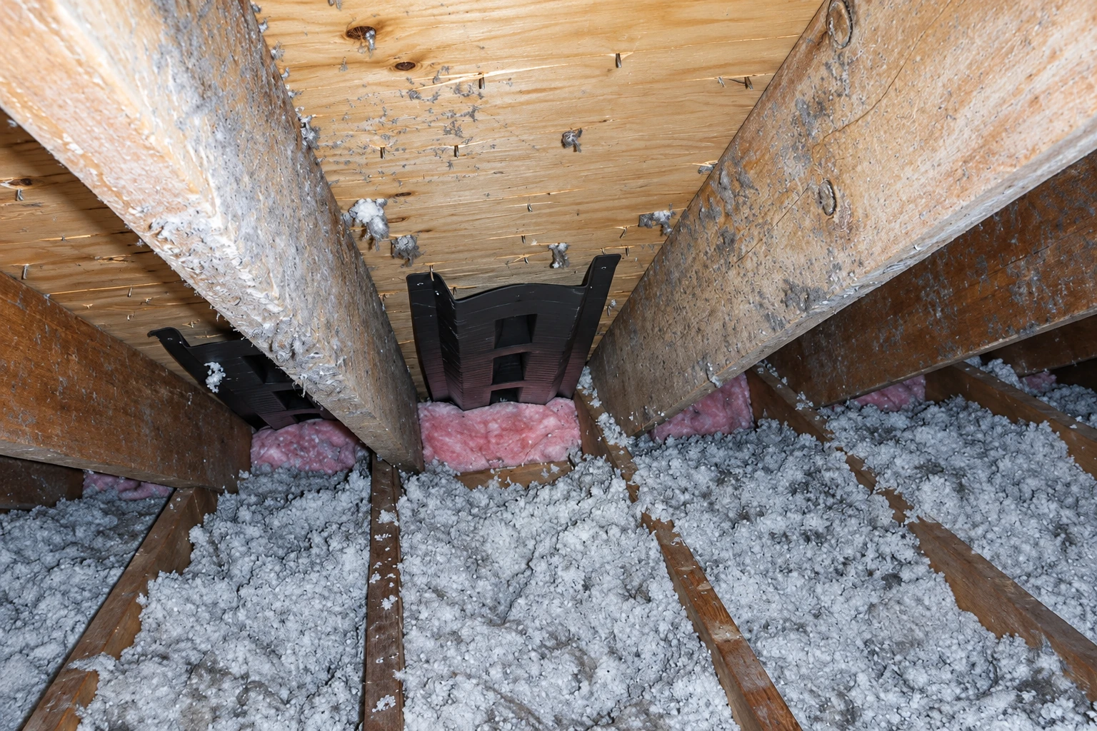 Loose-fill insulation covers an attic floor beneath exposed joists, with pink batts tucked between roof beams.