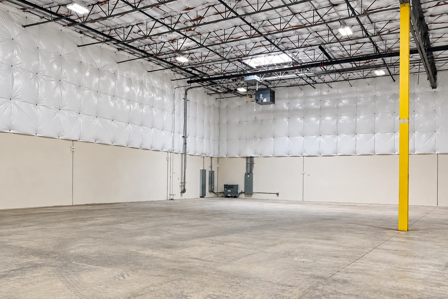 An empty commercial warehouse has reflective white insulation beneath its exposed roof trusses and a yellow support column.