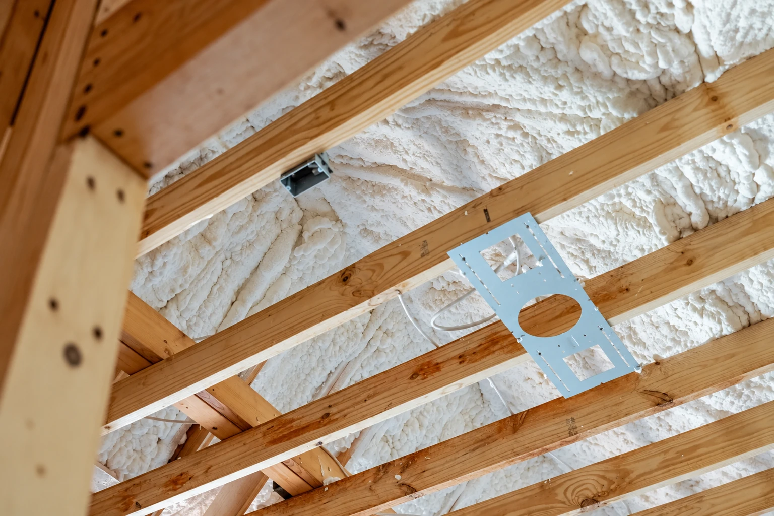 Open-cell spray foam fills the spaces between exposed wooden roof rafters, with a metal fixture plate hanging below.