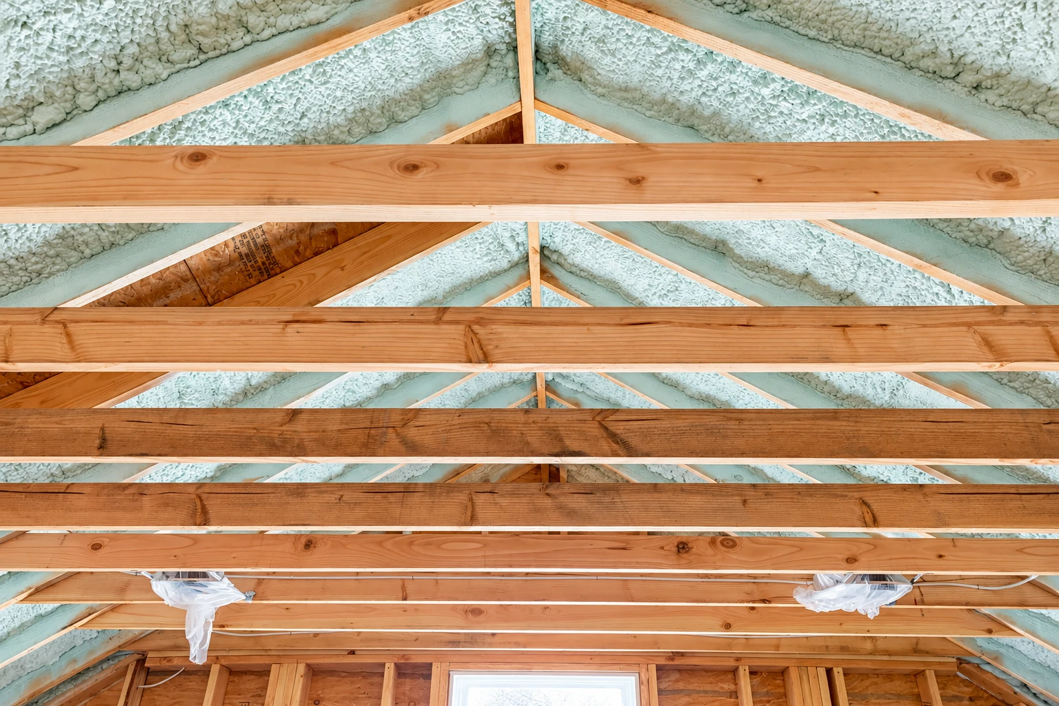 Spray foam insulation fills the spaces between exposed wooden roof trusses in an unfinished attic.