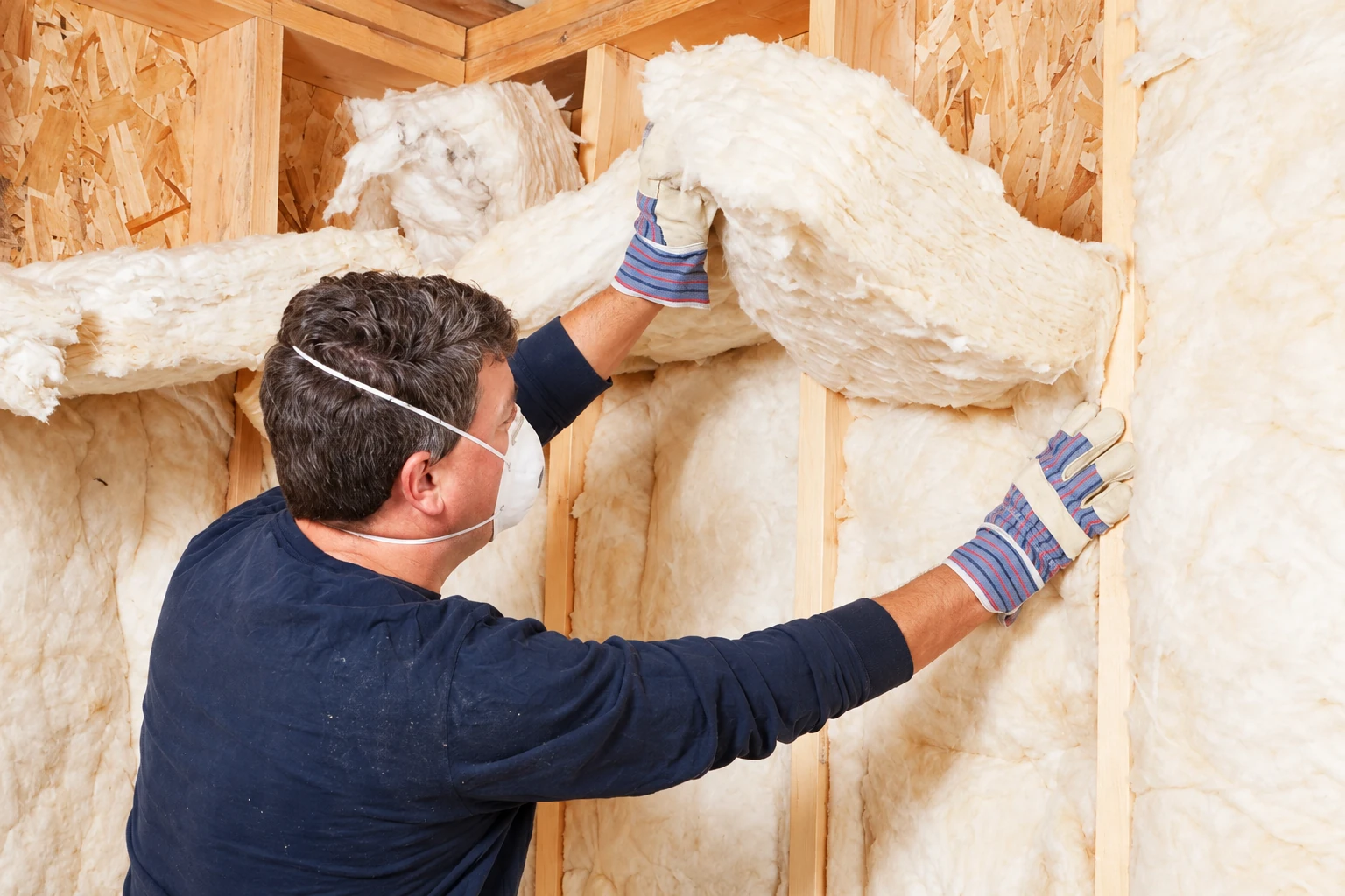 A worker wearing gloves and a mask fits fluffy white batt insulation between wall studs.
